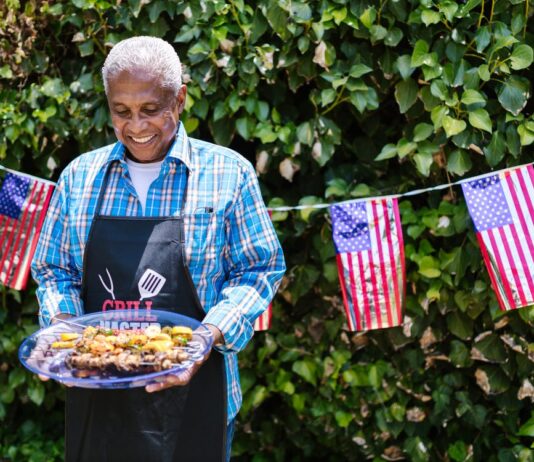 5 Memorial Day Gifts for Veterans and More Older adult with a plate full of fresh BBQ food against a hedge with American flags.