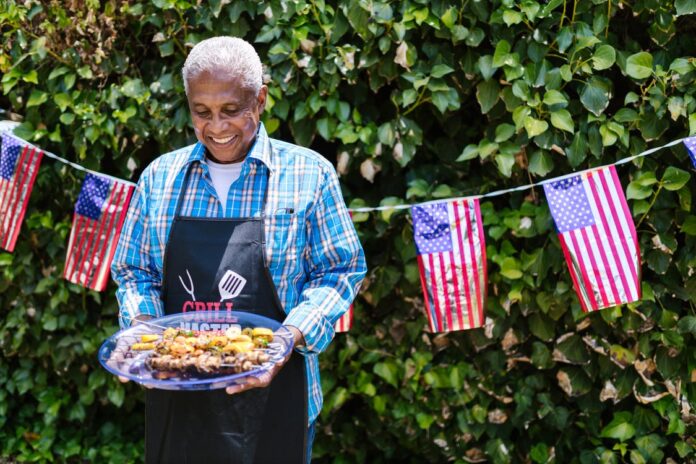Older adult with a plate full of fresh BBQ food against a hedge with American flags.