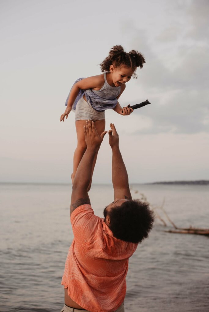 A young father is about to catch his young daughter on a beach. They are laughing and smiling.