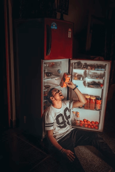 A tired college student sitting in front of an open fridge, eating snacks. 